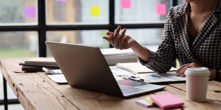 Frau mit Geste beim Planen einer Marketingstrategie, Laptop und Notizzettel im Hintergrund.